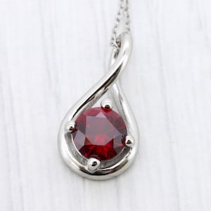 May include: A silver pendant necklace featuring a teardrop-shaped design. The pendant holds a round, deep red gemstone, secured by small prongs. The necklace chain is delicate and silver, creating a classic and elegant look.