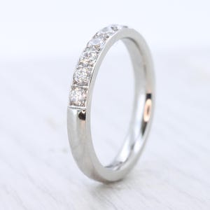 May include: A silver ring featuring a line of small, clear gemstones. The ring is displayed on a white surface, with a polished, reflective finish. This ring is a classic design, suitable for various occasions. The gemstones are set in a row.