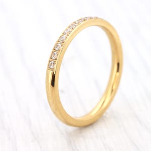 Peut inclure: Une bague en or avec une rangée de petits diamants carrés sur le dessus. La bague a un design simple et élégant, avec une finition polie. La bague est posée sur un fond blanc.