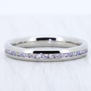 May include: A silver-colored ring with a channel setting of small, light purple and clear stones. The ring is round and has a polished finish, reflecting light. The gemstones are evenly spaced around the band, creating a continuous line of color.