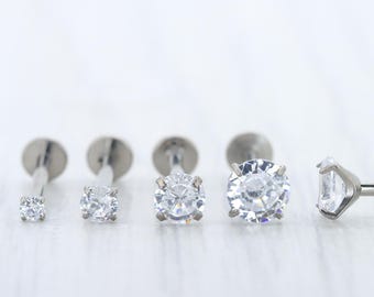 May include: An assortment of silver-toned, labret-style earrings with clear, sparkling cubic zirconia stones. The earrings vary in size, with the largest stone appearing to be approximately 6mm in diameter. The earrings are displayed on a white surface.