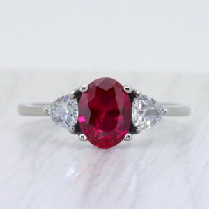 Könnte beinhalten: Ein silberner Ring mit einem großen, oval geschliffenen, tiefroten Edelstein, flankiert von zwei kleineren, birnenförmigen, klaren Edelsteinen. Der Ring hat ein dünnes, poliertes Band, und die Edelsteine sind in einer klassischen Krappenfassung gefasst.