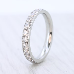 May include: A silver ring with a band of small, clear gemstones. The ring is set against a soft, white background. The gemstones are evenly spaced around the band, creating a continuous sparkle. This ring is a piece of jewelry.