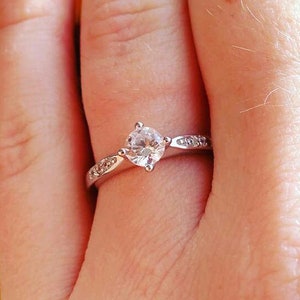 May include: A close-up of a hand wearing a silver ring with a round, clear gemstone. The ring is set with smaller gemstones on the band. The hand is holding a sunflower.
