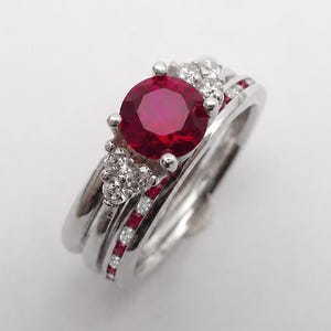 May include: A silver-toned ring set featuring a central round-cut red ruby flanked by small clear stones. The band includes a secondary ring component adorned with a channel of tiny red rubies and clear stones, creating a layered appearance.
