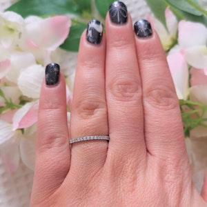May include: A silver ring with a row of small, clear gemstones. The ring is on a person's finger, with a floral background. The person's nails are painted with a dark color and have a decorative design.