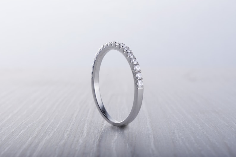 May include: A silver band ring with a row of small, clear gemstones set along the top. The ring is displayed on a light gray surface, with the focus on the sparkling stones and the polished metal.