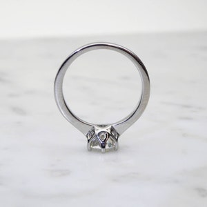May include: A silver engagement ring with a round, clear gemstone. The ring has a classic, solitaire design with a four-prong setting. The band is smooth and polished, reflecting light. The ring is set against a white marble background.
