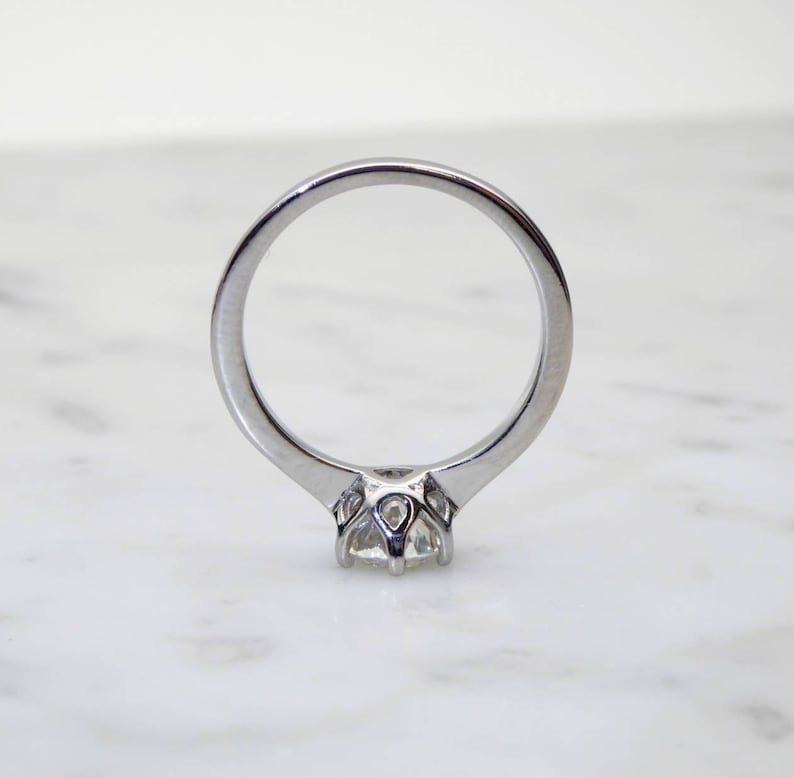 May include: A silver engagement ring with a round, clear gemstone. The ring has a classic, solitaire design with a four-prong setting. The band is smooth and polished, reflecting light. The ring is set against a white marble background.