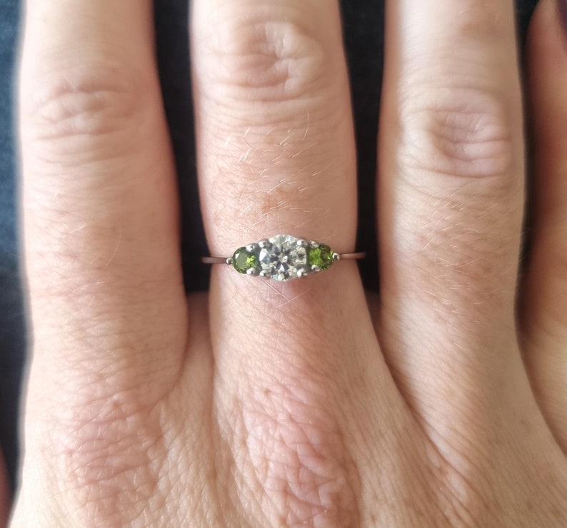 May include: A silver ring with a large, round, clear center stone and two smaller, round, green stones on either side.
