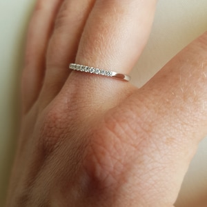 May include: A silver ring with a row of small, clear gemstones set along the top. The ring is on a finger, and the background is a soft, neutral color.