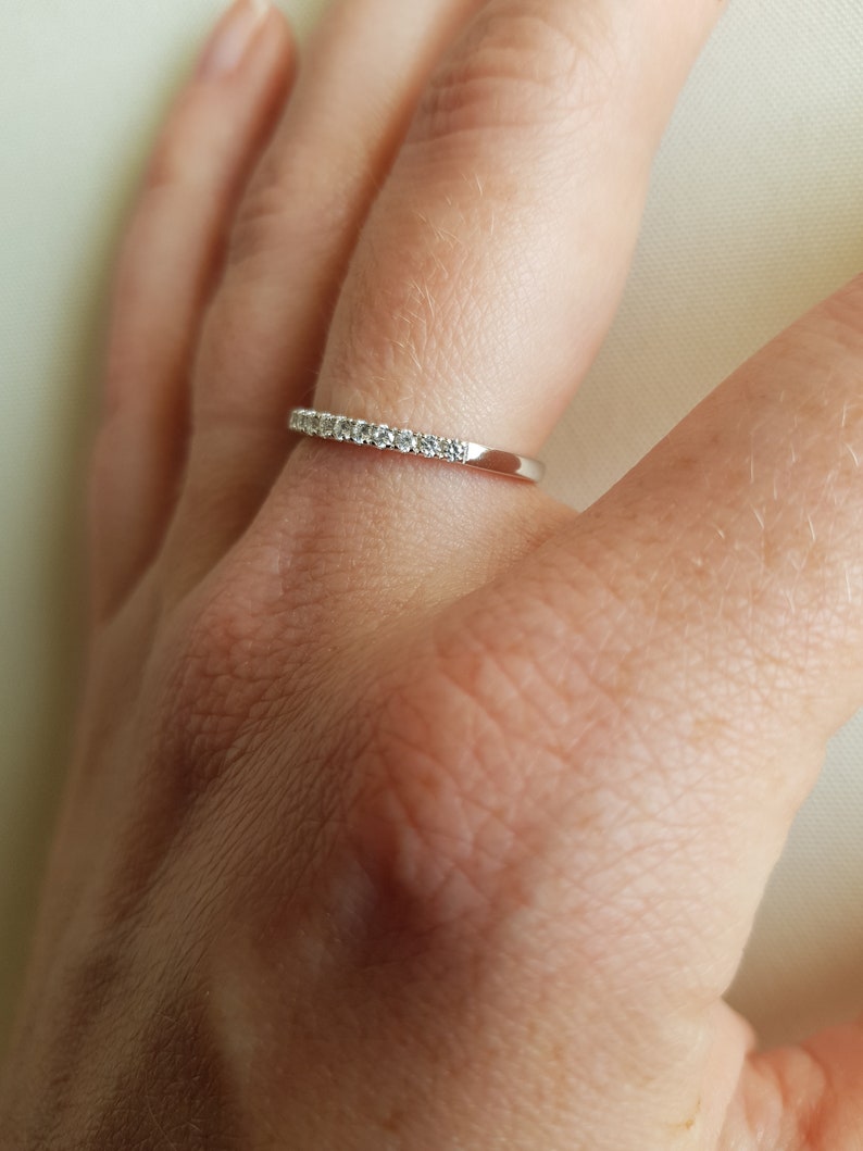 May include: A silver ring with a row of small, clear gemstones set along the top. The ring is on a finger, and the background is a soft, neutral color.