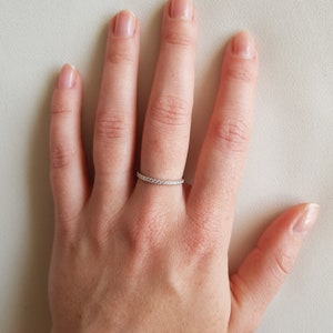 May include: A silver ring with a band of small, clear stones is worn on a person's finger. The ring is set against a neutral background. The ring appears to be a wedding band or eternity ring.