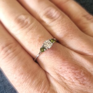 May include: A silver ring with a large, clear center stone and two smaller green stones on either side.