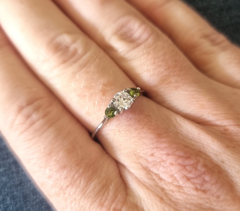 May include: A silver ring with a large, clear center stone and two smaller green stones on either side.