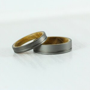 Puede incluir: Dos alianzas de boda, una con una banda de metal gris y una incrustaci&oacute;n de madera, y la otra con una banda de metal gris con una ranura.