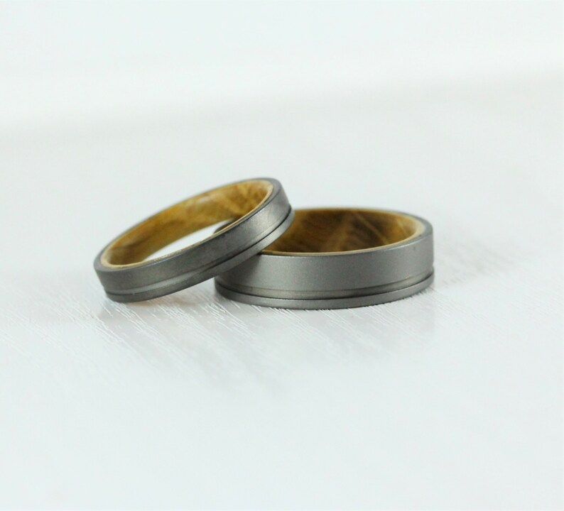 Puede incluir: Dos alianzas de boda, una con una banda de metal gris y una incrustaci&oacute;n de madera, y la otra con una banda de metal gris con una ranura.