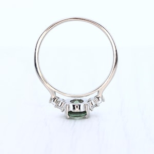 May include: A silver ring with a central oval-cut green gemstone, flanked by smaller, clear stones. The ring's band is thin and polished, with a delicate design around the main stone. The ring is set against a white background.