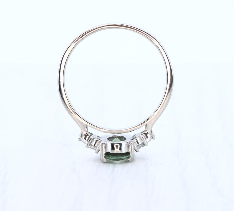 May include: A silver ring with a central oval-cut green gemstone, flanked by smaller, clear stones. The ring's band is thin and polished, with a delicate design around the main stone. The ring is set against a white background.