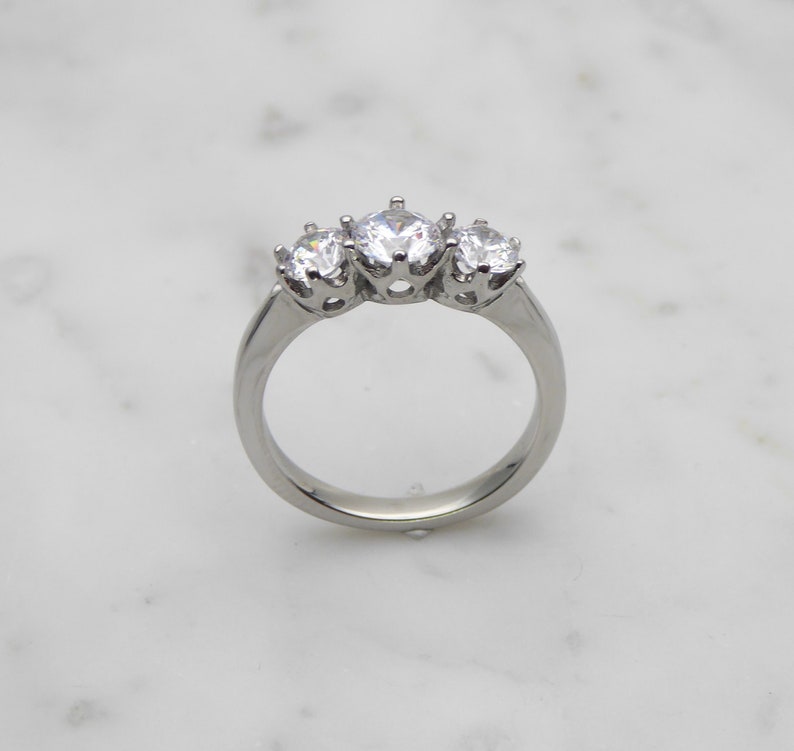 May include: A silver ring with three round clear stones set in prongs. The stones are arranged in a row, with the largest stone in the center.