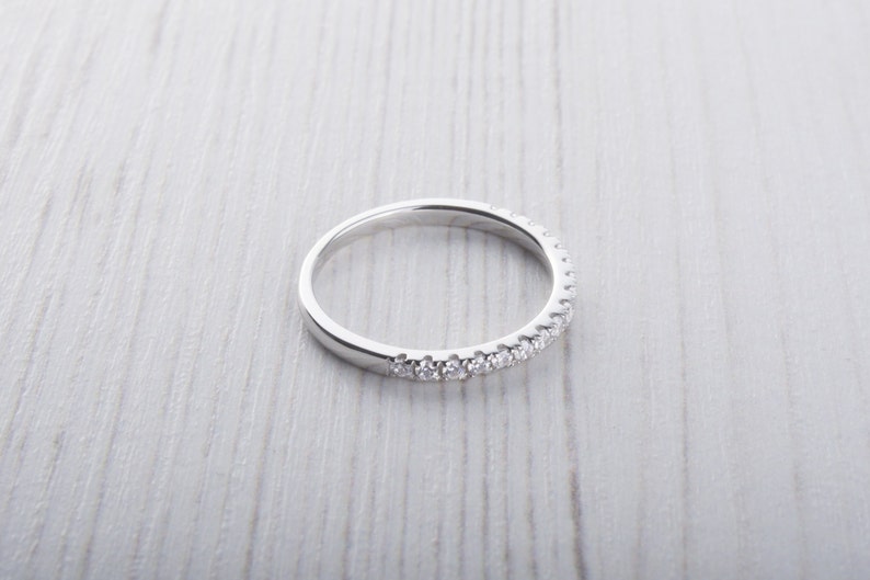 May include: A silver ring with a band of small, clear gemstones. The ring is set against a light-colored, textured surface. The gemstones are evenly spaced around the top half of the ring.