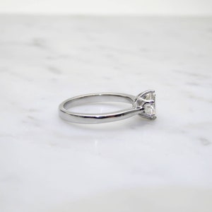 May include: A silver engagement ring with a large, clear, square-cut gemstone. The ring is set on a white marble surface, showcasing the polished metal and the brilliance of the stone. The ring's band is smooth and rounded.