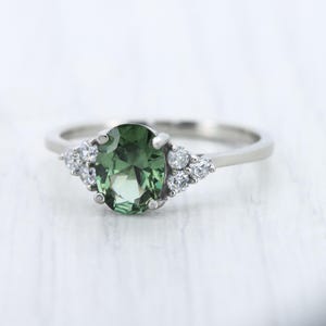 May include: A silver ring featuring a large, oval-cut green gemstone as the centerpiece, flanked by clusters of smaller, clear gemstones. The ring's band is a simple, polished silver.