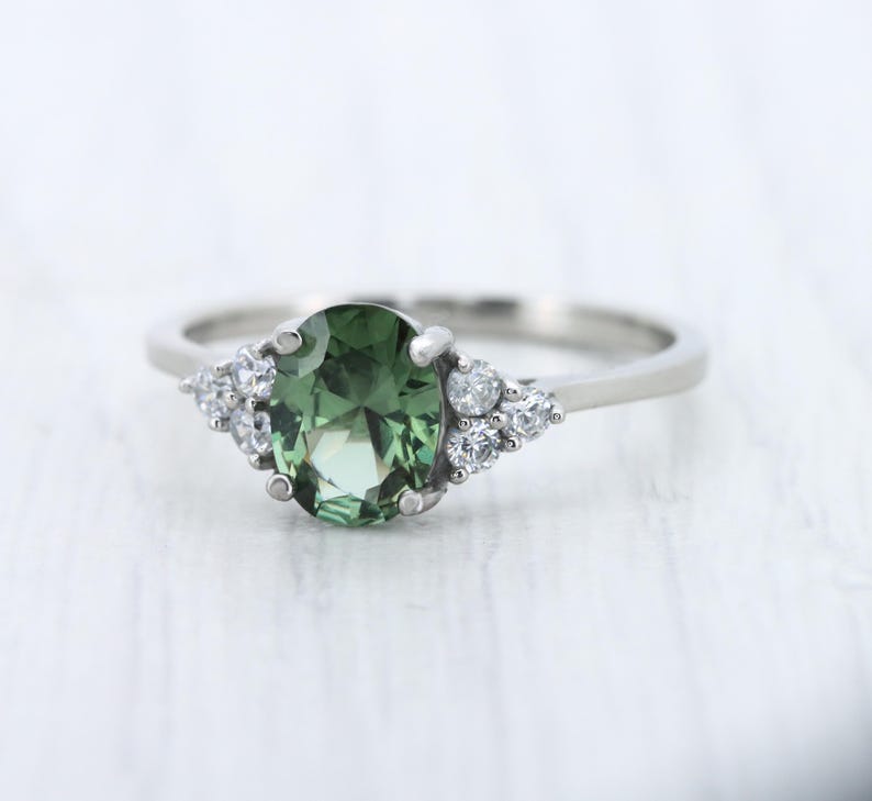 May include: A silver ring featuring a large, oval-cut green gemstone as the centerpiece, flanked by clusters of smaller, clear gemstones. The ring's band is a simple, polished silver.