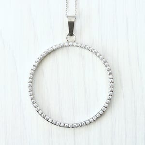 Puede incluir: Un collar plateado con un colgante circular. El colgante es un anillo de pequeñas piedras preciosas transparentes. El collar cuelga de una fina cadena de plata, creando un diseño sencillo y elegante.