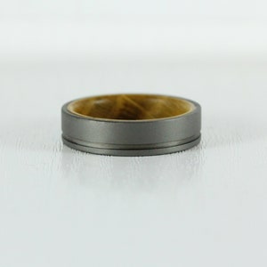Puede incluir: Un anillo de titanio gris con una incrustaci&oacute;n de madera. El anillo tiene un borde biselado y un acabado pulido.
