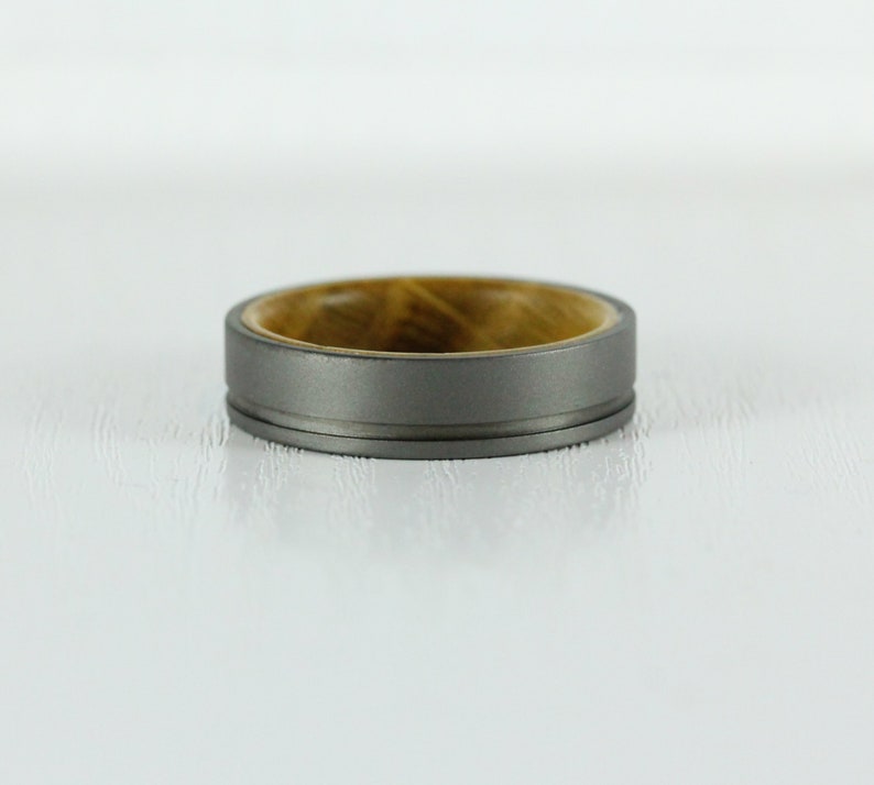 Puede incluir: Un anillo de titanio gris con una incrustaci&oacute;n de madera. El anillo tiene un borde biselado y un acabado pulido.