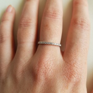 May include: A delicate silver ring with a band of small, clear gemstones. The ring is worn on a finger, showcasing its simple yet elegant design. The gemstones sparkle, catching the light.