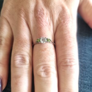 May include: A silver ring with a large, round, clear center stone and two smaller, round, green stones on either side.