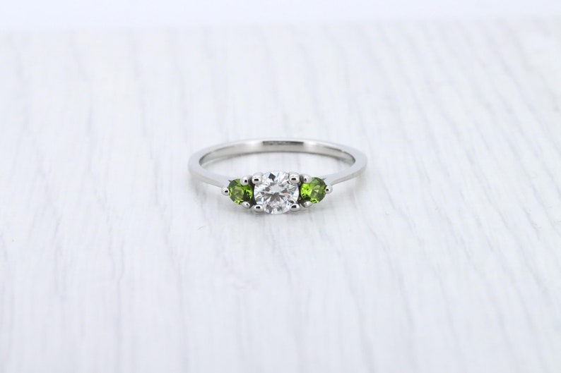 May include: A silver ring with a round, clear center stone and two smaller, round, green stones on either side.