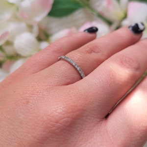 May include: A delicate silver ring with a continuous row of small, clear gemstones. The ring is set on a finger, with a soft-focus floral background. The ring's design is simple and elegant, suitable for everyday wear or special occasions.