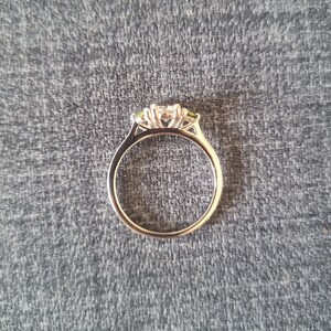 May include: A silver ring with a three-stone setting. The center stone is a large, clear diamond, and the two side stones are smaller, green gemstones.
