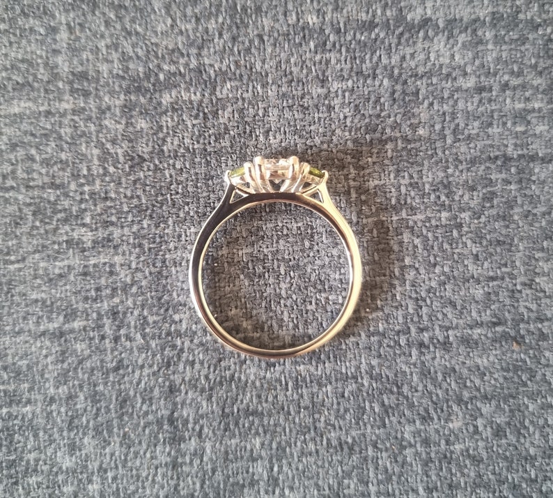 May include: A silver ring with a three-stone setting. The center stone is a large, clear diamond, and the two side stones are smaller, green gemstones.
