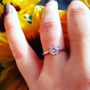 May include: A silver engagement ring with a large, round, clear gemstone. The ring is set on a hand with a sunflower in the background. The ring's band is thin and simple.
