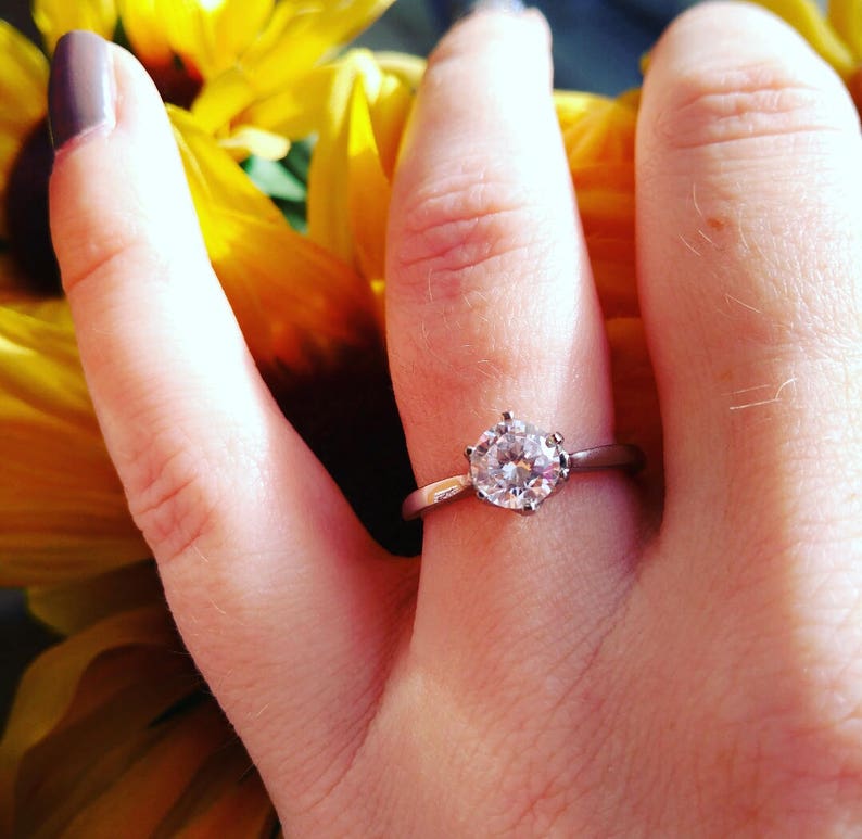 May include: A silver engagement ring with a large, round, clear gemstone. The ring is set on a hand with a sunflower in the background. The ring's band is thin and simple.