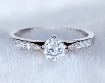 May include: A silver ring with a large, round, clear gemstone as the centerpiece. The band is also adorned with smaller, clear gemstones. The ring is set against a soft, white background.