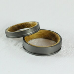 Puede incluir: Dos anillos de boda con un exterior de metal gris y una incrustaci&oacute;n de madera. Los anillos est&aacute;n apilados uno encima del otro.