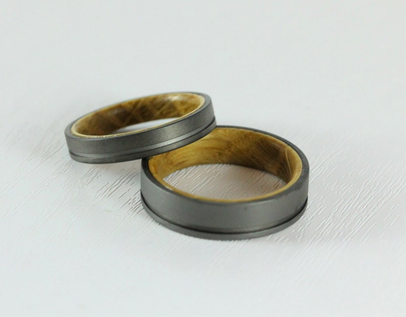 Puede incluir: Dos anillos de boda con un exterior de metal gris y una incrustaci&oacute;n de madera. Los anillos est&aacute;n apilados uno encima del otro.
