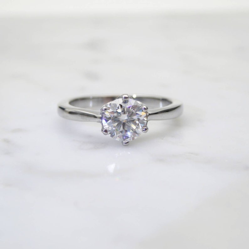 May include: A silver engagement ring with a large, round, clear gemstone. The gemstone is held in place by six prongs. The ring is set against a white marble surface.