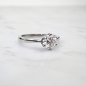 May include: A silver engagement ring with a large, round, clear gemstone. The ring has a classic six-prong setting, and the band is smooth and polished. The ring is set against a white marble background.