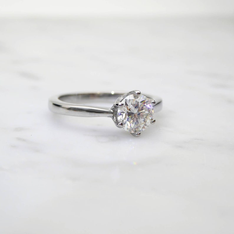 May include: A silver engagement ring with a large, round, clear gemstone. The ring has a classic six-prong setting, and the band is smooth and polished. The ring is set against a white marble background.