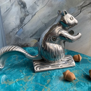 May include: A silver squirrel nutcracker with a detailed design. The squirrel is holding a nut in its paws and has a long, curved tail that acts as a lever. There are three acorns on a blue surface.