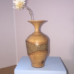 May include: A wooden vase with a brown and beige patterned design, sitting on a light blue shelf. A single white flower is in the vase.