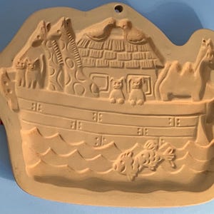 May include: A tan ceramic mould showing Noah's Ark, featuring pairs of animals like giraffes, camels, and birds. The ark has a detailed roof and windows, with waves at the base. A hole is at the top for hanging.
