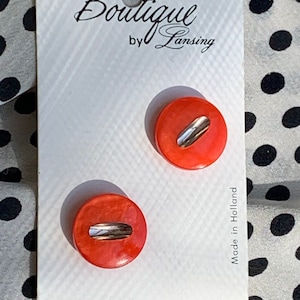 May include: Two orange plastic buttons with a silver stripe across the centre. The buttons are on a white card with the text "Boutique by Lansing", "$ 1.30", "28-3/4", "2 on card", "9977", "5997", and "Made in Holland".