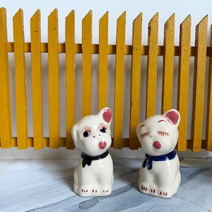 May include: Two white ceramic dog salt and pepper shakers. The salt shaker has a black bow tie and the pepper shaker has a blue bow tie. Both have pink ears and painted details on their faces.